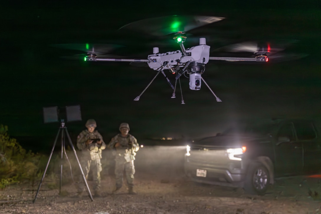 A drone flies low as two soldiers in camouflage uniforms stand by a tripod and watch, one holding a controller, with headlights of a parked truck partly lighting the darkness.