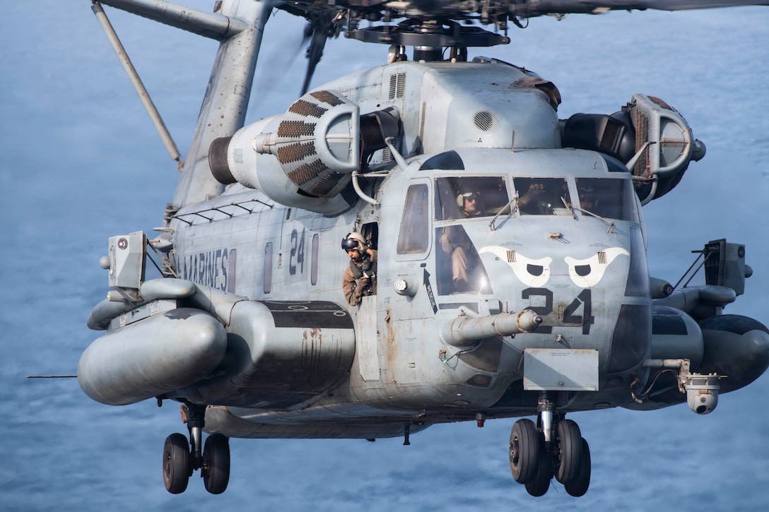 A Marine wearing a flight suit and flight helmet looks out the door of a helicopter in flight over a body of water on a bright day.