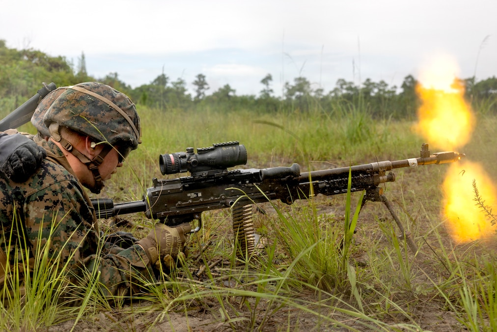 A Marine wearing a camouflage uniform and helmet fires a machine gun from a prone position in a grassy field during daytime.
