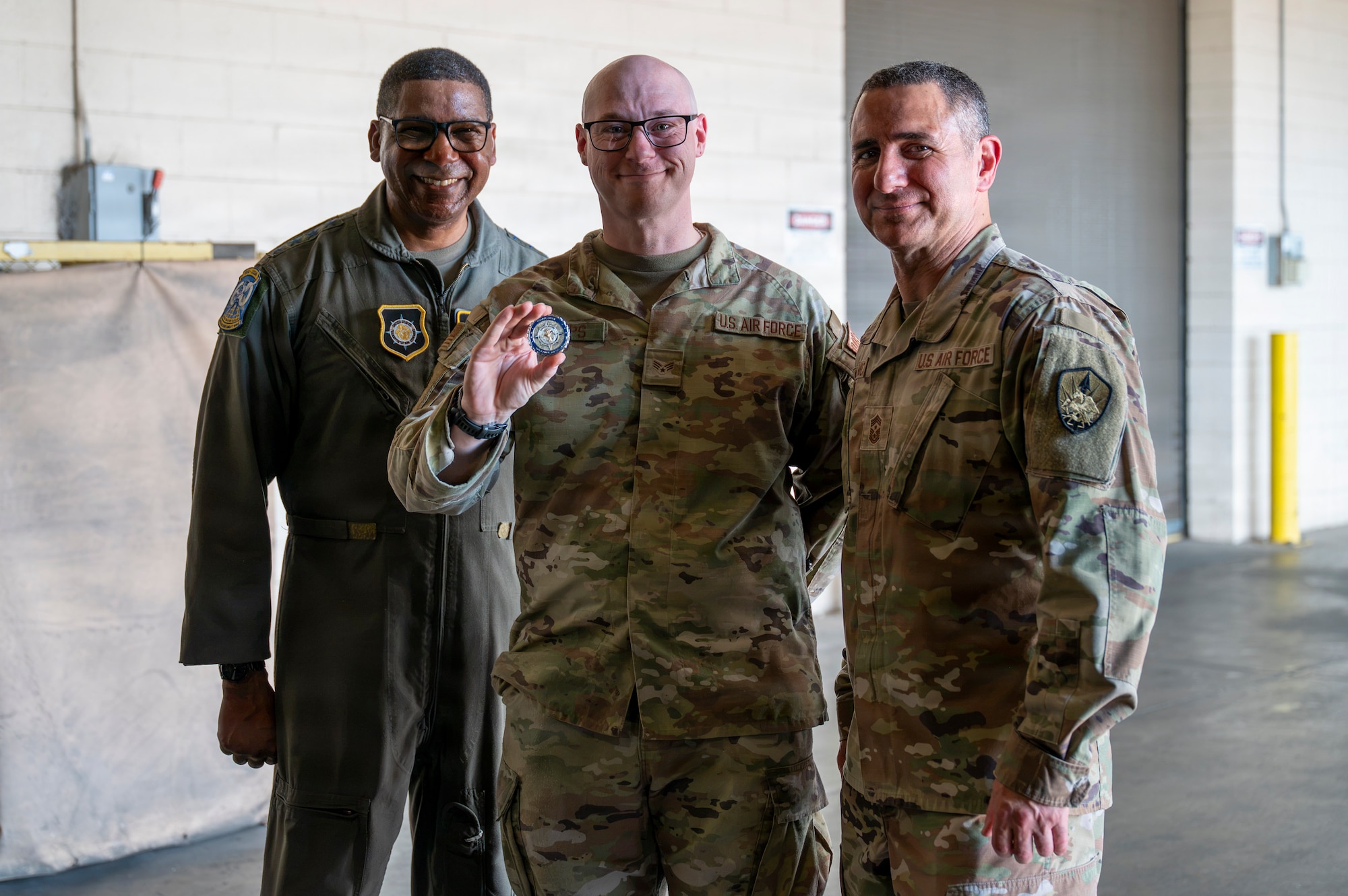 Three smiling people stand in a warehouse, the person in the middle holds out a coin.