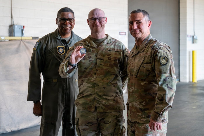 Three smiling people stand in a warehouse, the person in the middle holds out a coin.