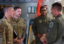 Four people stand in a group inside of an aircraft, three look towards the left of the image as one person speaks.