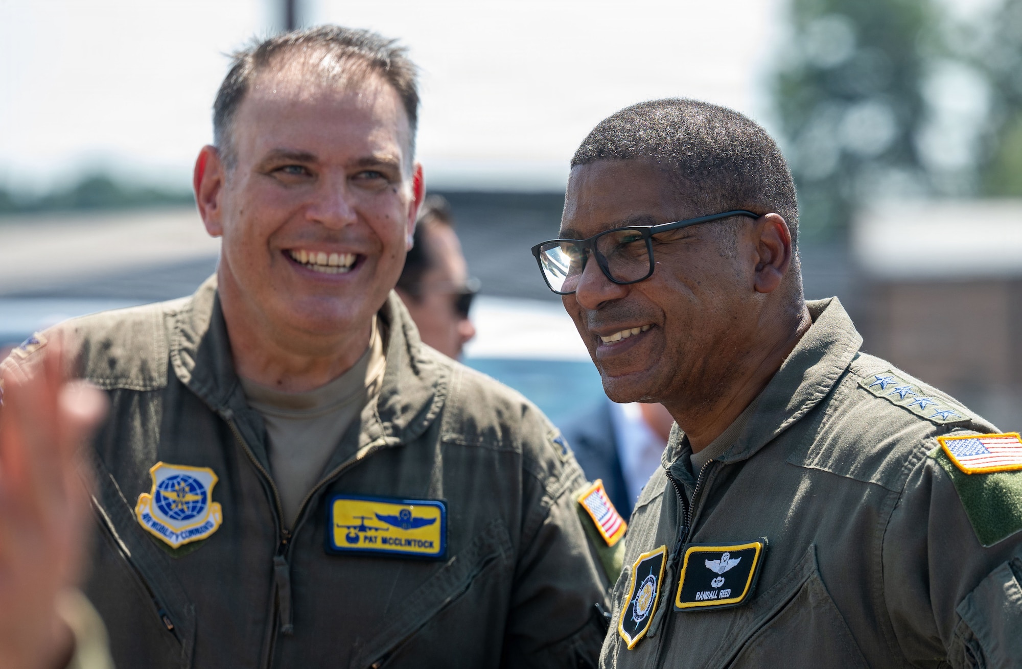 Two men smile, both wearing green uniforms with colorful patches.