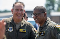 Two men smile, both wearing green uniforms with colorful patches.
