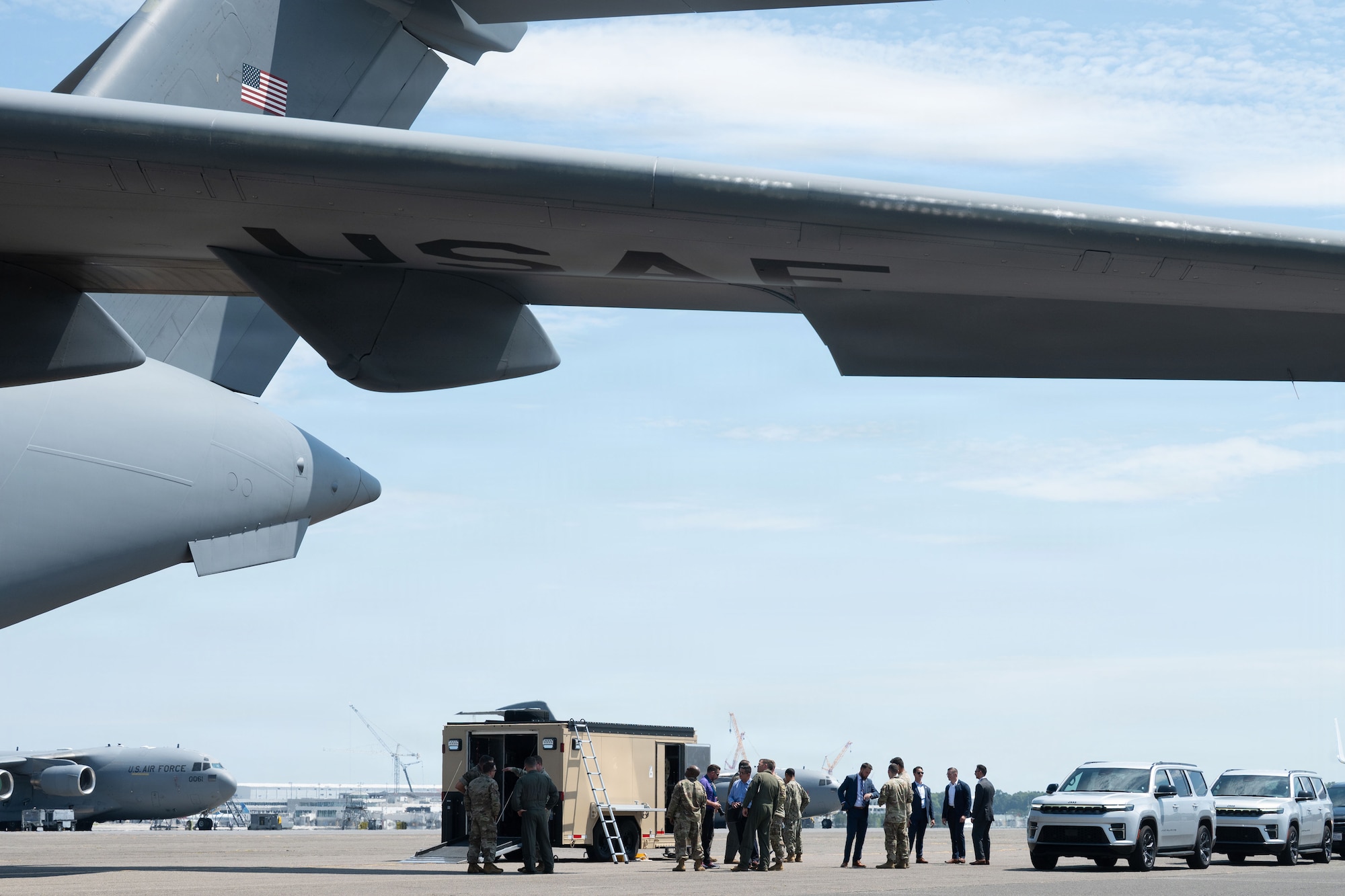A group of people standing next to a tan trailer and severa vehicles stand under the wing of a C-17 Globemaster III on the flightline.