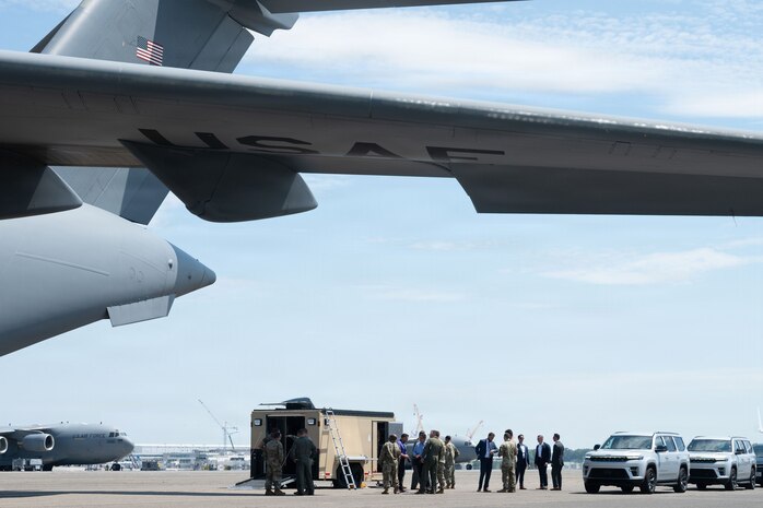 A group of people standing next to a tan trailer and severa vehicles stand under the wing of a C-17 Globemaster III on the flightline.
