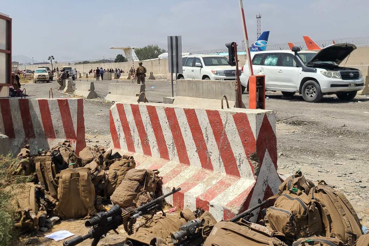Backpacks and weapons sit on the ground near a vehicle barrier on the side of a street. Cars and people are in the background.