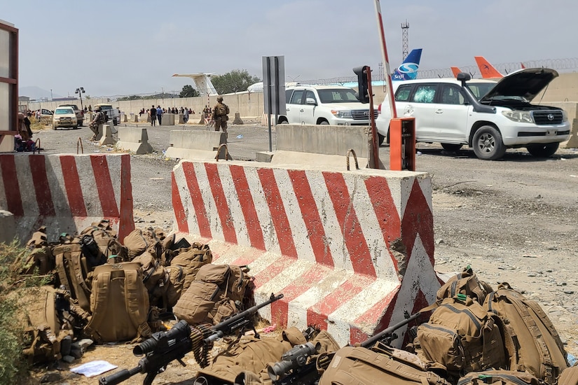 Backpacks and weapons sit on the ground near a vehicle barrier on the side of a street. Cars and people are in the background.
