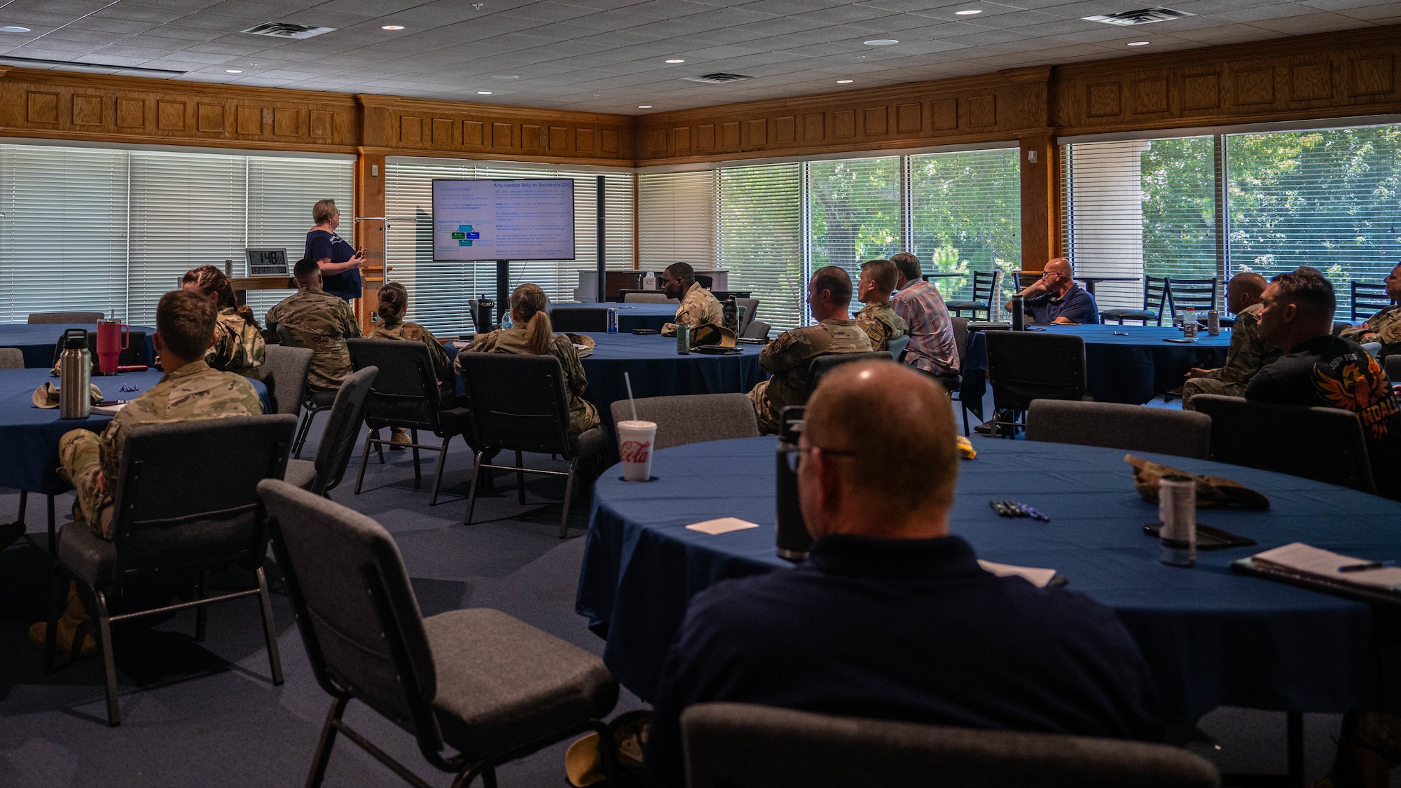 Civilians and military personnel in uniform sit in a room with tables covered in blue tablecloths.