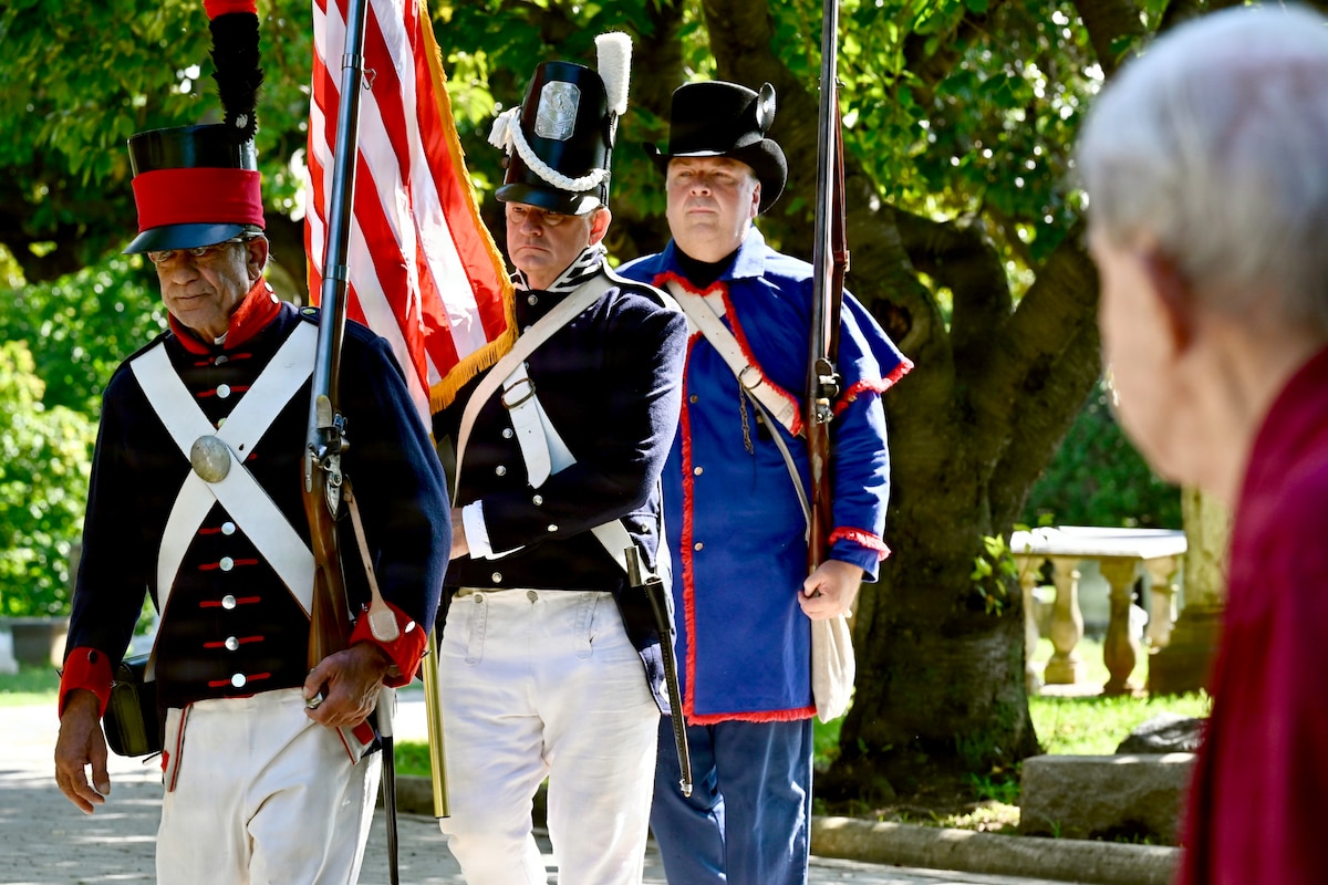 Three men dress in historical military uniforms stand in a line; the man in the middle is holding the American flag while the other two men are holding muskets.