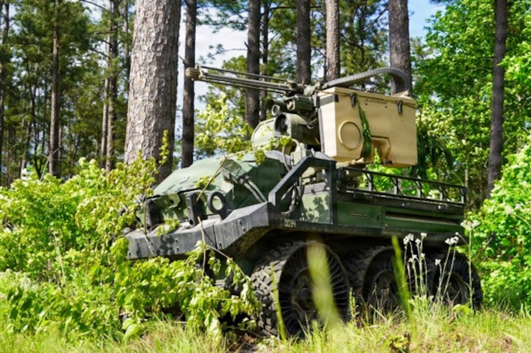 An unmanned ground vehicle with a large mounted gun is parked in a wooded area during daytime.