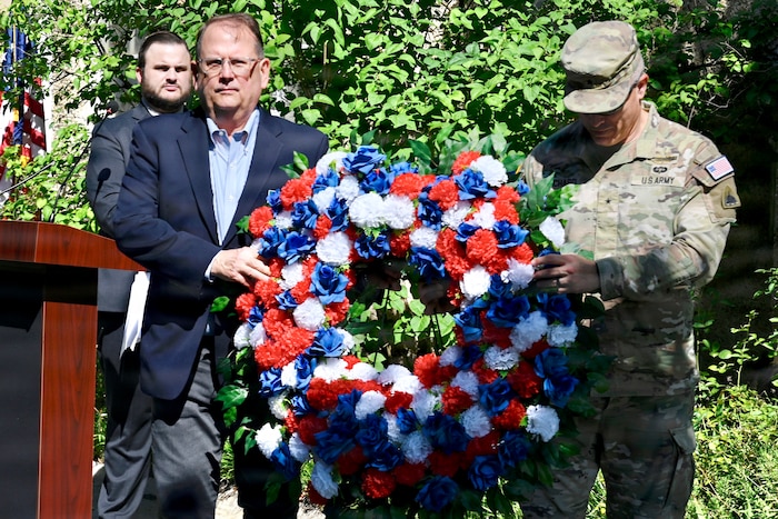 Two men, one in a camouflage military uniform and one in business attire, carry a large wreath made of red, white and blue flowers.