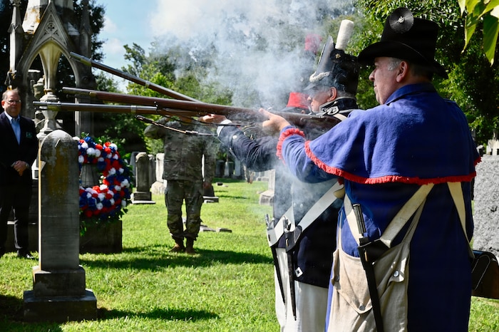 Three men in historic military uniforms fire rifles at a cemetery while smoke billows around them.