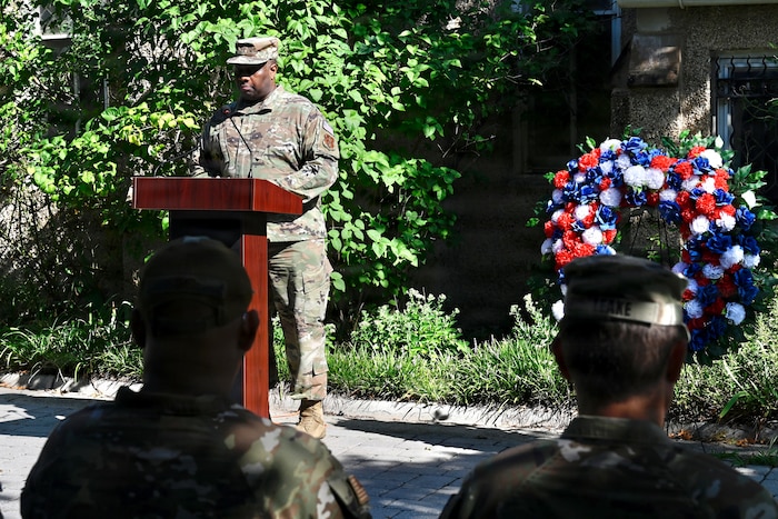 A man in a camouflage military uniform stands at a lectern to address a crowd seated outside.