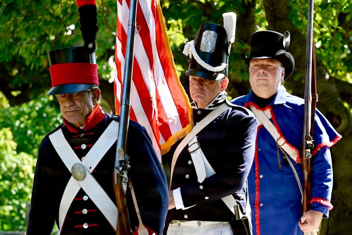 Three men dress in historical military uniforms stand in a line; the man in the middle is holding the American flag while the other two men are holding muskets.