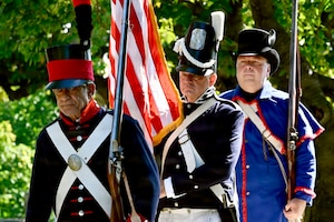 Three men dress in historical military uniforms stand in a line; the man in the middle is holding the American flag while the other two men are holding muskets.
