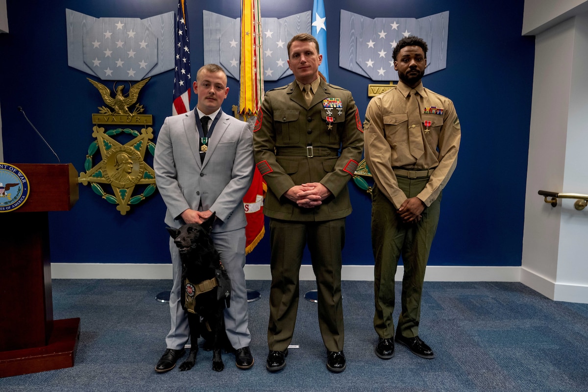 Three men, one wearing a blue business suit and the two others in military dress uniforms, stand in a straight line with flags behind them and three star medals topped with light blue ribbons on a blue wall behind the flags.