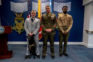 Three men, one wearing a blue business suit and the two others in military dress uniforms, stand in a straight line with flags behind them and three star medals topped with light blue ribbons on a blue wall behind the flags.