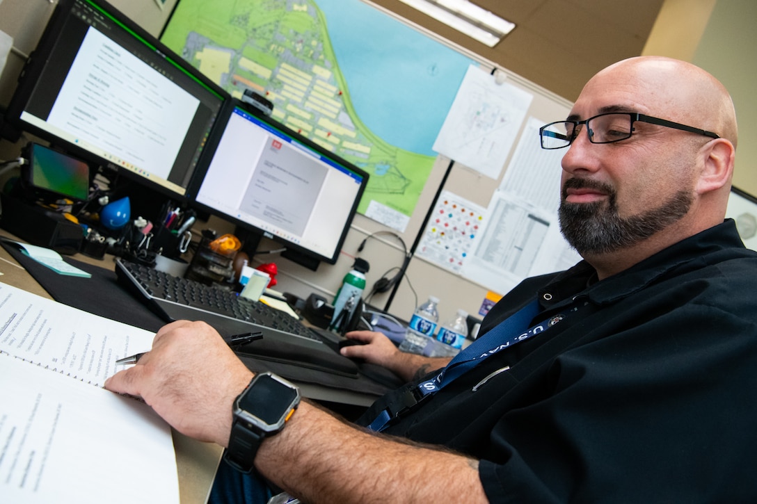 A man sits at a desk in front of a computer looking down at papers on the desk