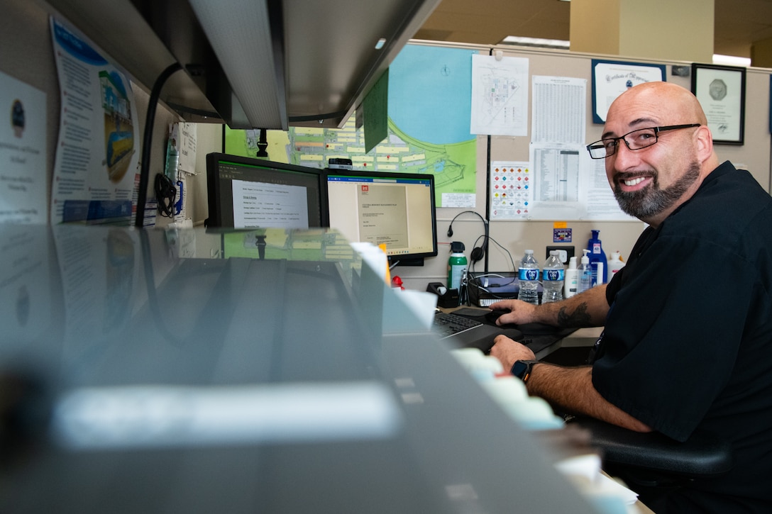 A man sits at a desk in front of a computer looking into the camera and smiling