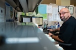 A man sits at a desk in front of a computer looking into the camera and smiling