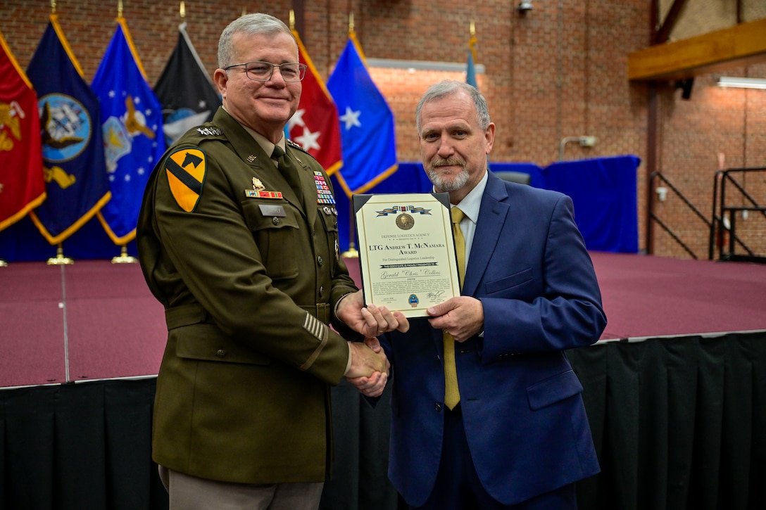 A man in a olive green Army u8niform smiles as he shakes the hand of another man in a suit in front of an open stage in a large multipurpose room.