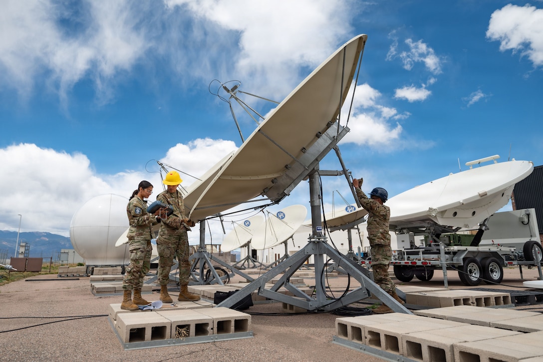A Guardian manipulates a large satellite dish outdoors as two others stand nearby, one holding a rectangular device.