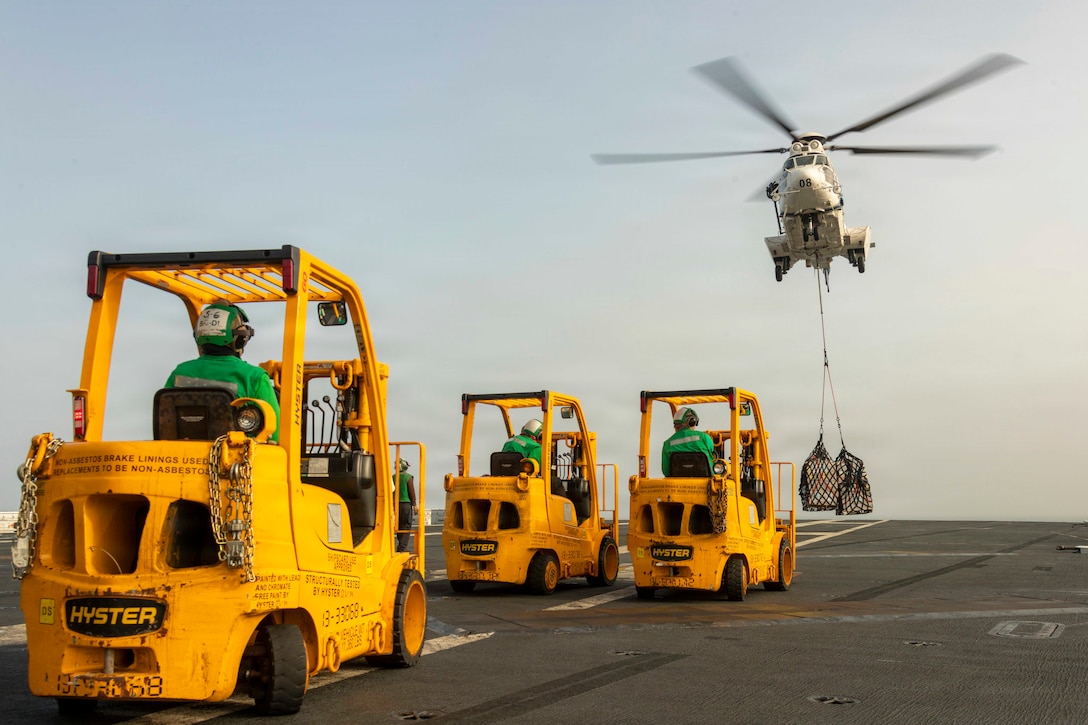 A helicopter carrying netted cargo hovers over a ship’s flight deck as sailors in green gear sit in three yellow forklifts during the day.