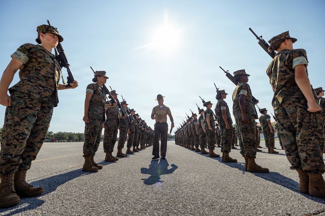 A Marine walks in the center of recruits standing in rows in formation holding a weapon on their shoulder during the day.