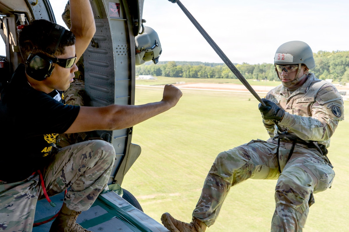 A soldier wearing camouflage pants, a black T-shirt, and a communications headset signals with a clenched fist to another soldier wearing a camouflage uniform, a helmet, and a harness holding a rope as he hangs out of a helicopter hovering over a field on a bright day.