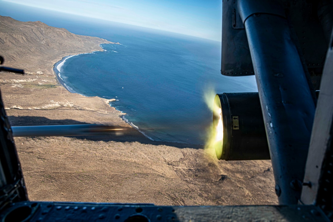 A missile shoots out of a launcher mounted to a helicopter flying above a coastline on a bright day.