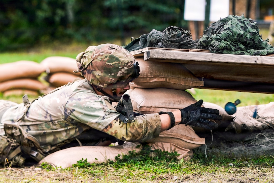 A soldier wearing a camouflage uniform, helmet and load carrying equipment throws a grenade into a bunker from a prone position on a bright day.