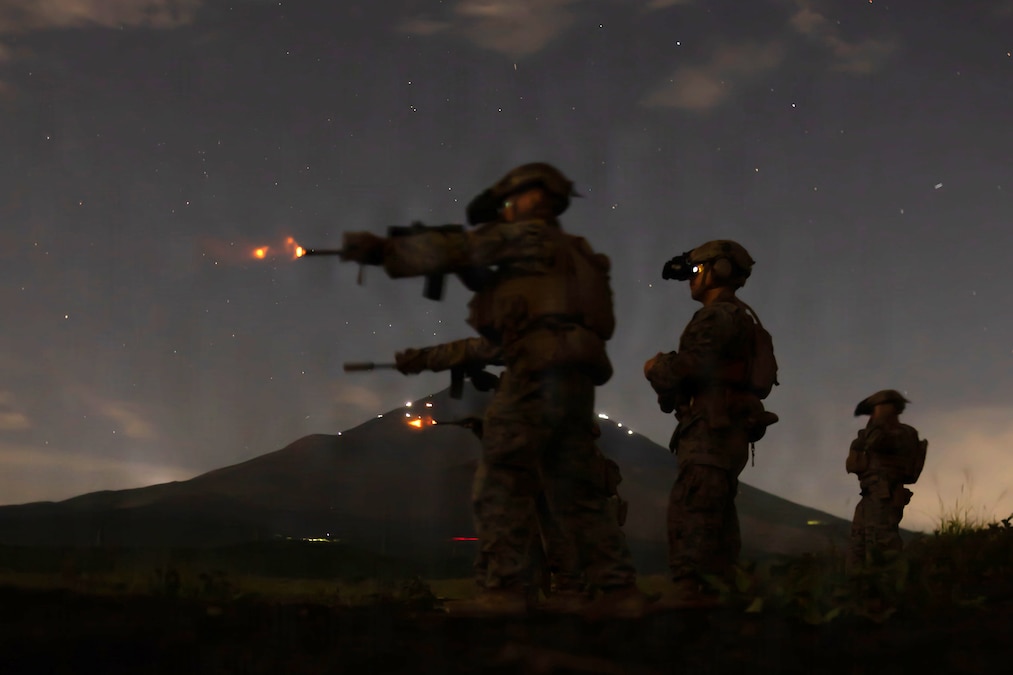 Three Marines fire rifles from a standing position as two other Marines observe them with all of them wearing helmets and night vision equipment on a dark night.