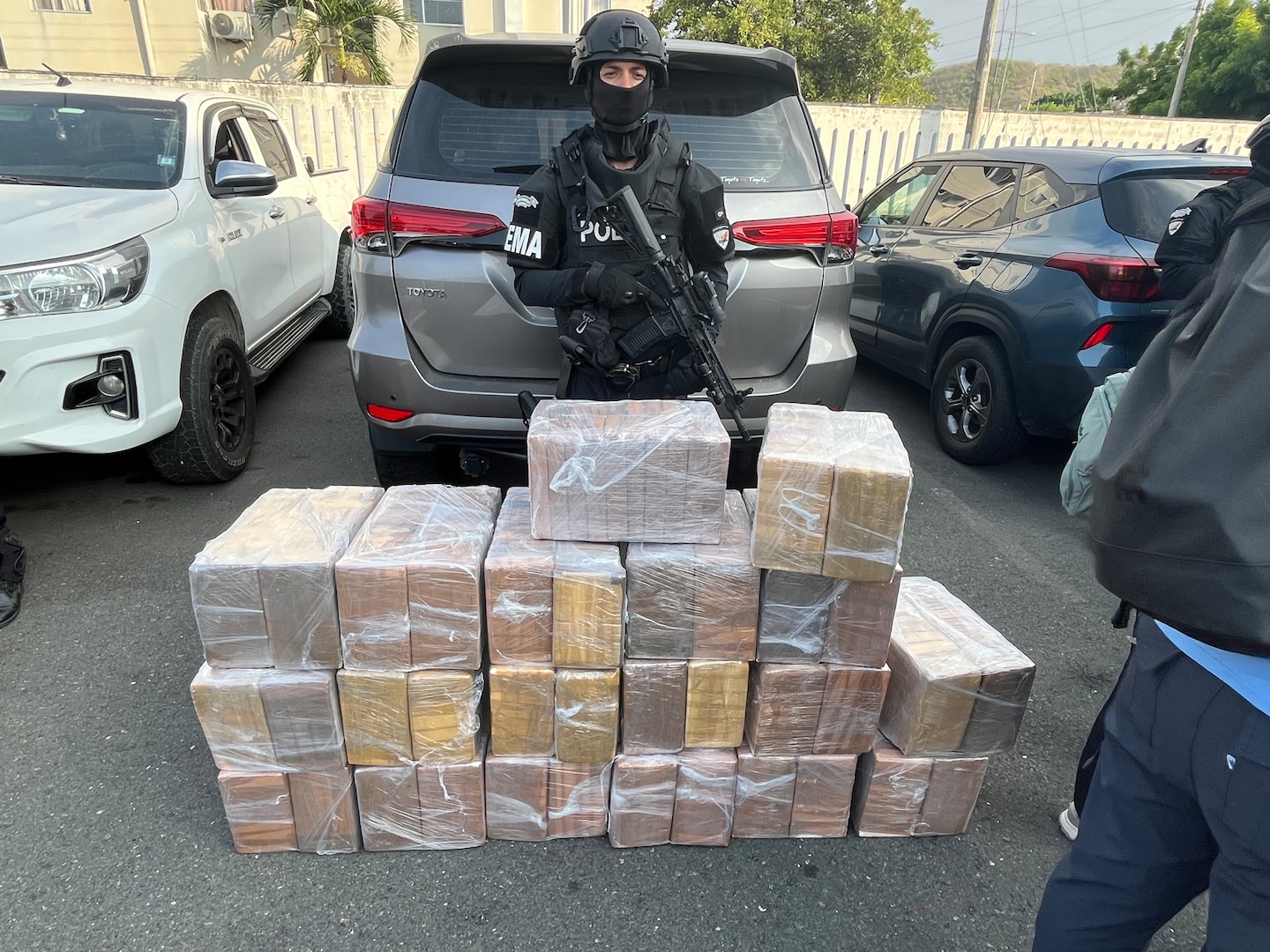 A masked officer holding a rifle stands behind a stack of seized illicit drugs.