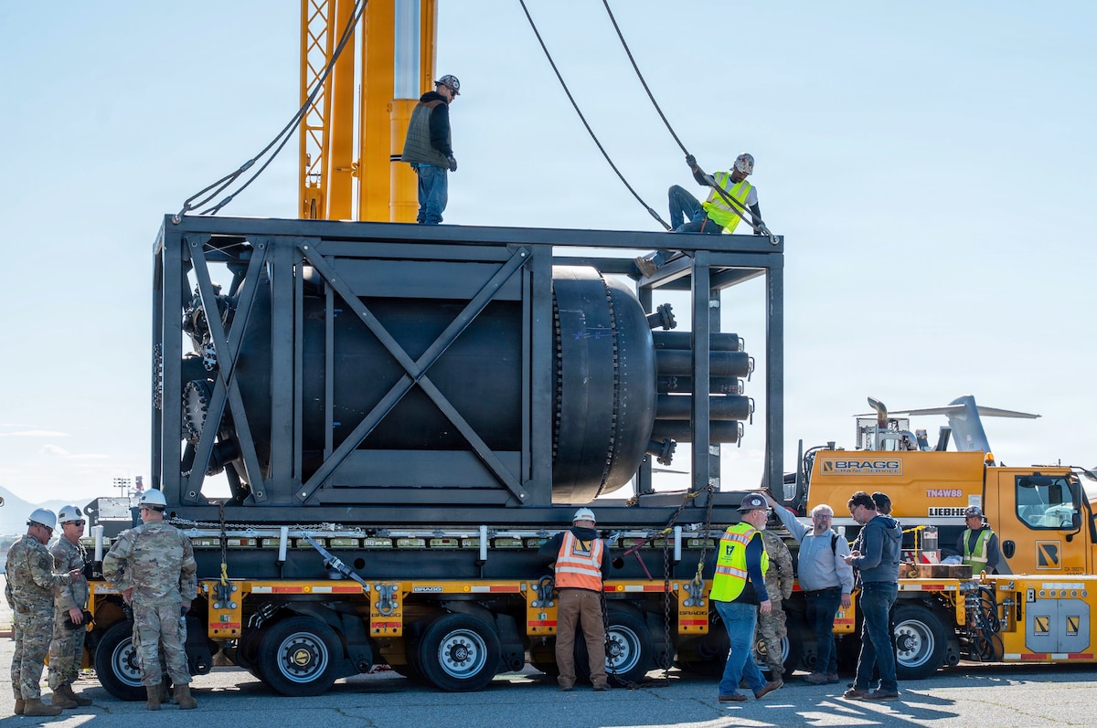 Two men in casual attire and hard hats attach cables to a steel structure sitting on the back of a trailer while several other people in similar attire and camouflage military uniforms work on the trailer.