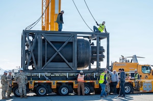 Two men in casual attire and hard hats attach cables to a steel structure sitting on the back of a trailer while several other people in similar attire and camouflage military uniforms work on the trailer.