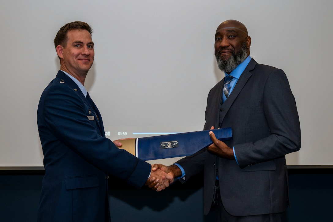 2 men shake hands and hold an award.