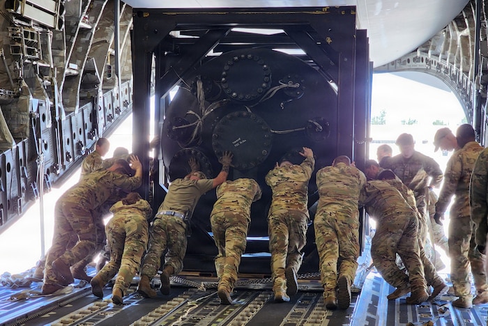 A dozen people in military camouflage uniforms push a nuclear reactor out of the loading bay of a military aircraft.