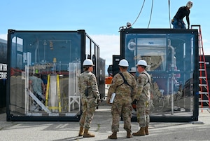 Three people in camouflage military uniforms and hard hats stand in front of two large clear containers while another man in casual attire and a hard hat works on top one of the containers.