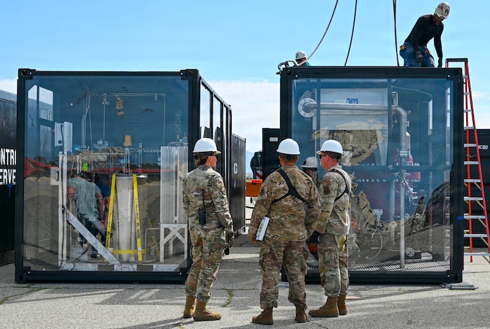 Three people in camouflage military uniforms and hard hats stand in front of two large clear containers while another man in casual attire and a hard hat works on top one of the containers.