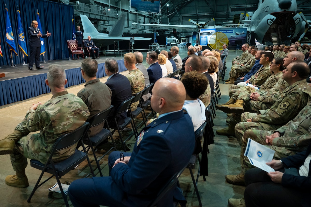 People in crowd watching man in Air Force dress blues speak on stage behind a podium.