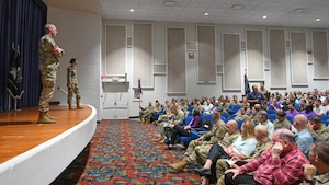 U.S. Space Force Col. Charles Cooper and Chief Master Sgt. Sarah Williams stand on a stage as they listen to a member of the audience speak.