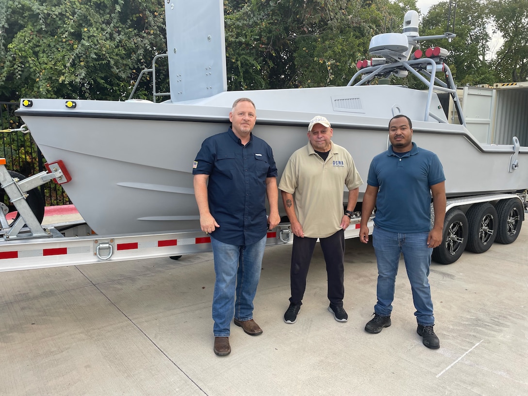 Three men stand in front of a drone vessel on a trailer