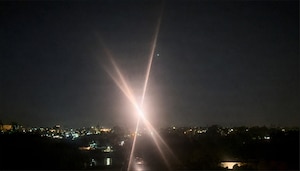 A bright light emits flares into the night sky over a city.