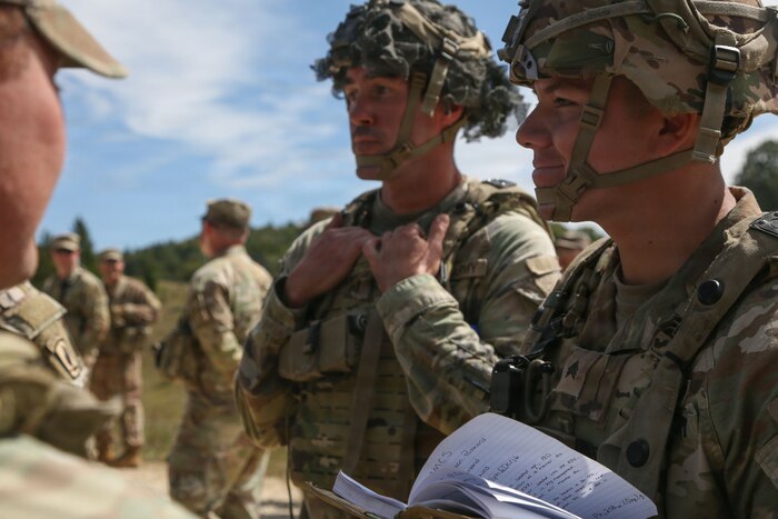 A close up of two men wearing combat gear, one is holding a notepad.