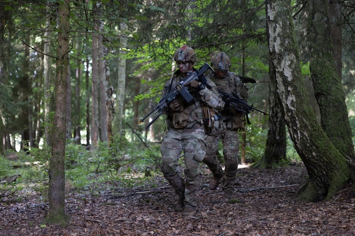 Two people wearing military combat gear walk through a forest.