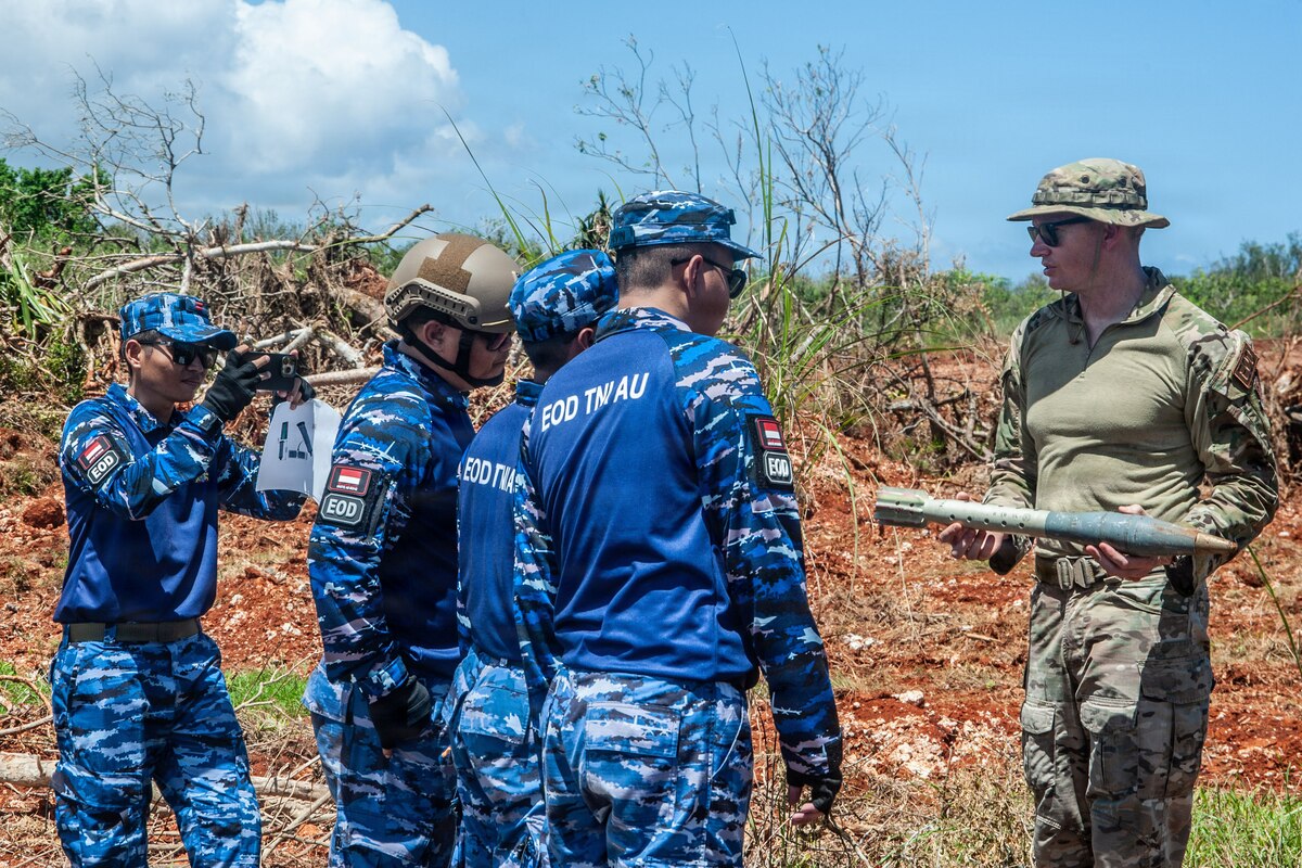 A man in a camouflage military uniform holds a military weapon outside in a grassy terrain while talking with a group of people in similar attire.