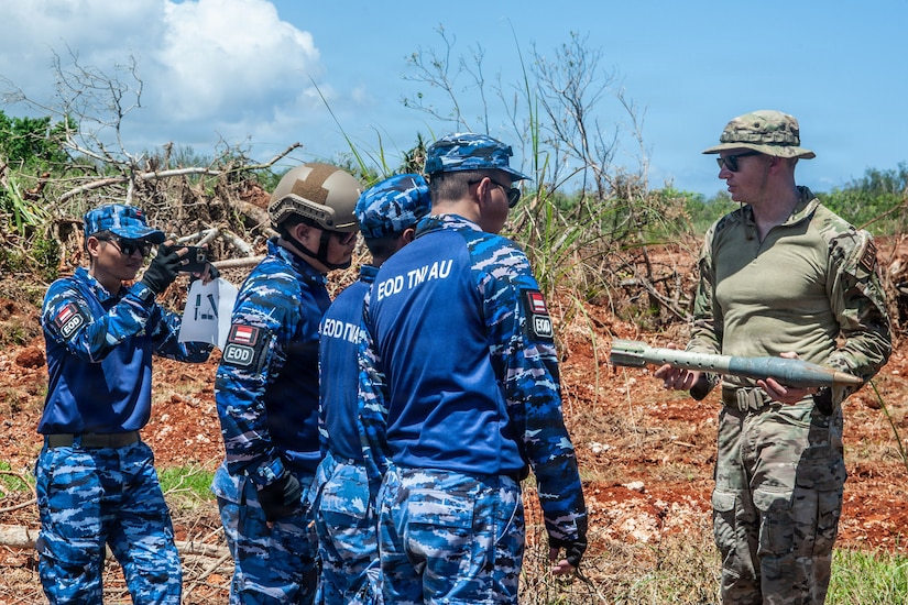A man in a camouflage military uniform holds a military weapon outside in a grassy terrain while talking with a group of people in similar attire.