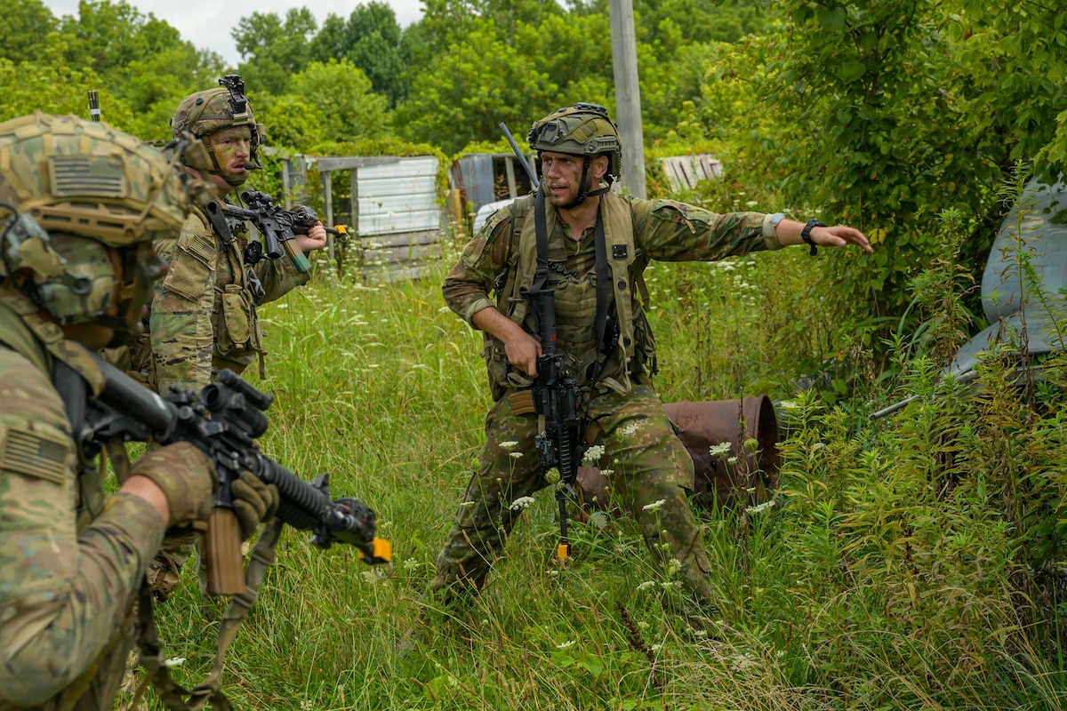 A man in a camouflage military uniform and helmet carries a weapon as he leads two other people in similar attire through a wooded area with tall grass.