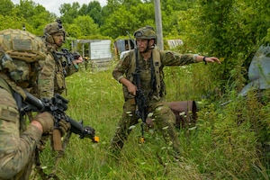 A man in a camouflage military uniform and helmet carries a weapon as he leads two other people in similar attire through a wooded area with tall grass.
